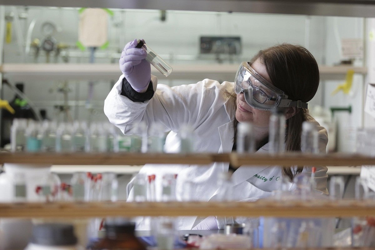 Image of a professor in a lab coat with protective eyewear looking at contents in a glass test tube